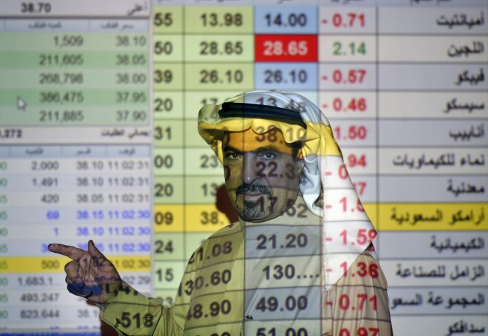 FILE- In this Thursday, Dec. 12, 2019, file photo, a trader talks to others in front of a screen displaying Saudi stock market values at the Arab National Bank in Riyadh, Saudi Arabia. Saudi Arabian oil company Aramco's initial public offering raised $29.4 billion, more than previously announced after the company said Sunday it used a so-called 